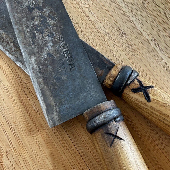 Close-up of two hand-forged kitchen knives with dark patina steel blades and wooden handles, shown on a bamboo surface to highlight artisan craftsmanship and aged metal texture.