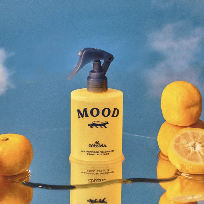 Mood all-purpose fragrance spray bottle styled with fresh lemons on a glossy blue reflective surface, bright citrus-inspired home scent product photography with a clean modern aesthetic.